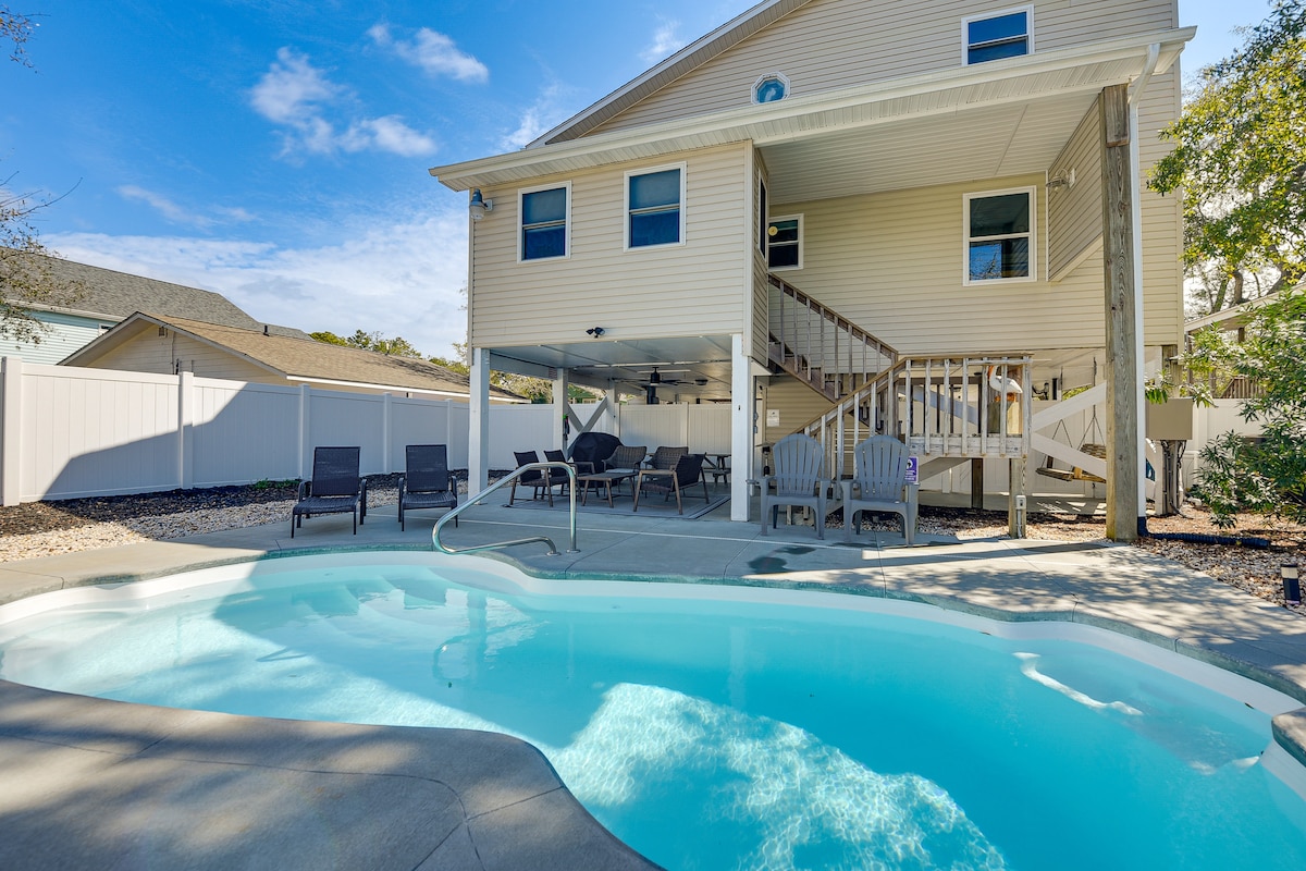 The outdoor area features a private, inviting pool with a gently sloping entrance, surrounded by a concrete deck. Several lounge chairs are positioned nearby, and a staircase leads to the elevated house above, showcasing a covered patio below.