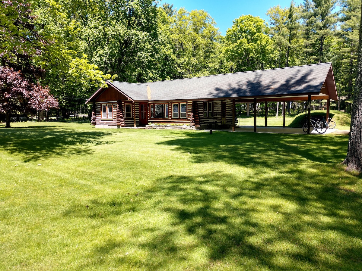 A traditional log cabin is set within a verdant lawn, surrounded by trees. The spacious front porch is shaded, and a bicycle is visible to the side. Natural light filters through the trees, creating dappled patterns on the grass.