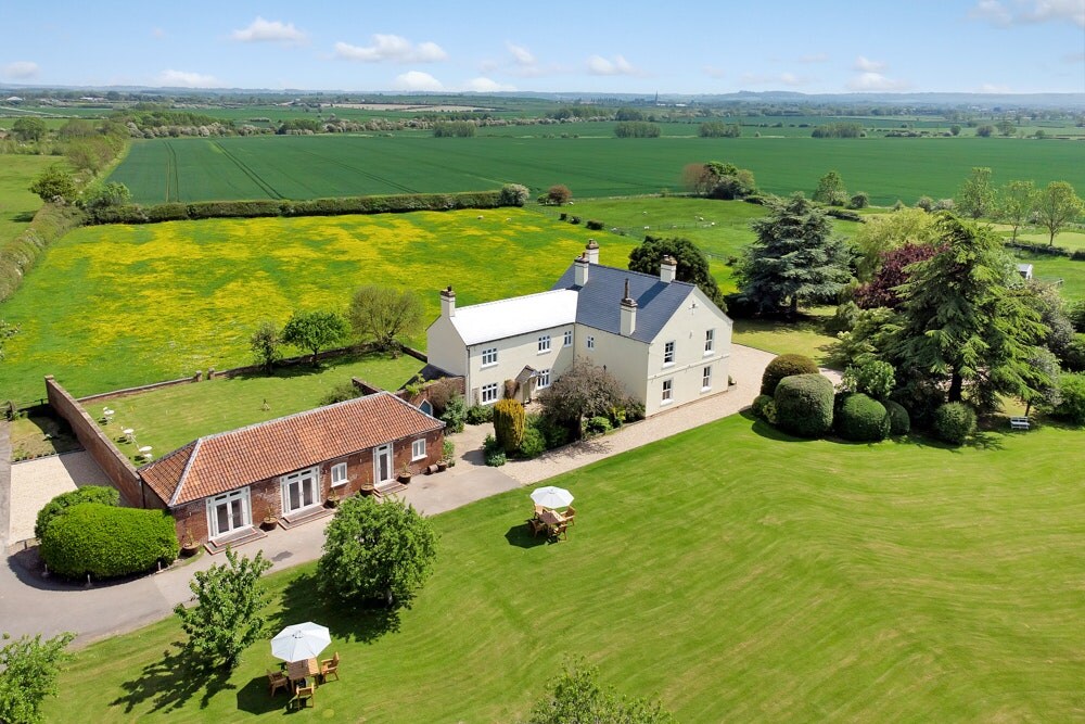 An aerial view showcases the Chestnuts Country House surrounded by expansive green fields and vibrant yellow patches of wildflowers. The main house features a light-colored exterior, while adjacent outbuildings are visible. Tables with umbrellas are placed on the lawn, inviting relaxation in the scenic outdoors.