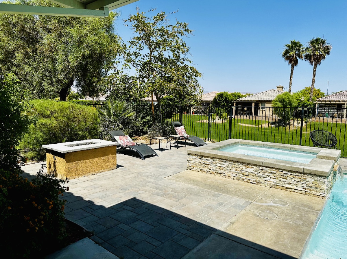 A private outdoor space features a hot tub and a neatly laid stone patio. Lounge chairs are positioned for relaxation, surrounded by lush greenery and palm trees. A protective fence encloses the area, ensuring privacy and a tranquil setting.