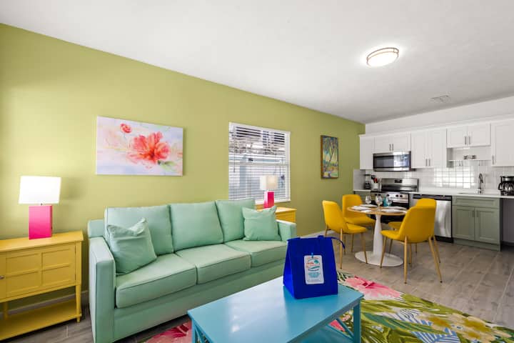 საცხოვრებელი და სასადილო სივრცე Siesta Key Beach‑ზე, სადაც პასტელის მწვანე დივანი, ყვითელი სკამები და ყვავილებიანი აქცენტები სანაპიროს მხარის ხასიათს გადმოსცემს