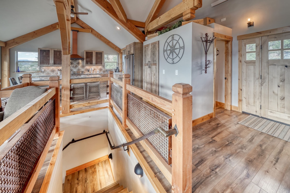An open staircase leads to the lower level, surrounded by wood railings and accented by natural light. The entryway features double doors, and the living area is visible in the background, showcasing timber beams and warm wooden flooring.