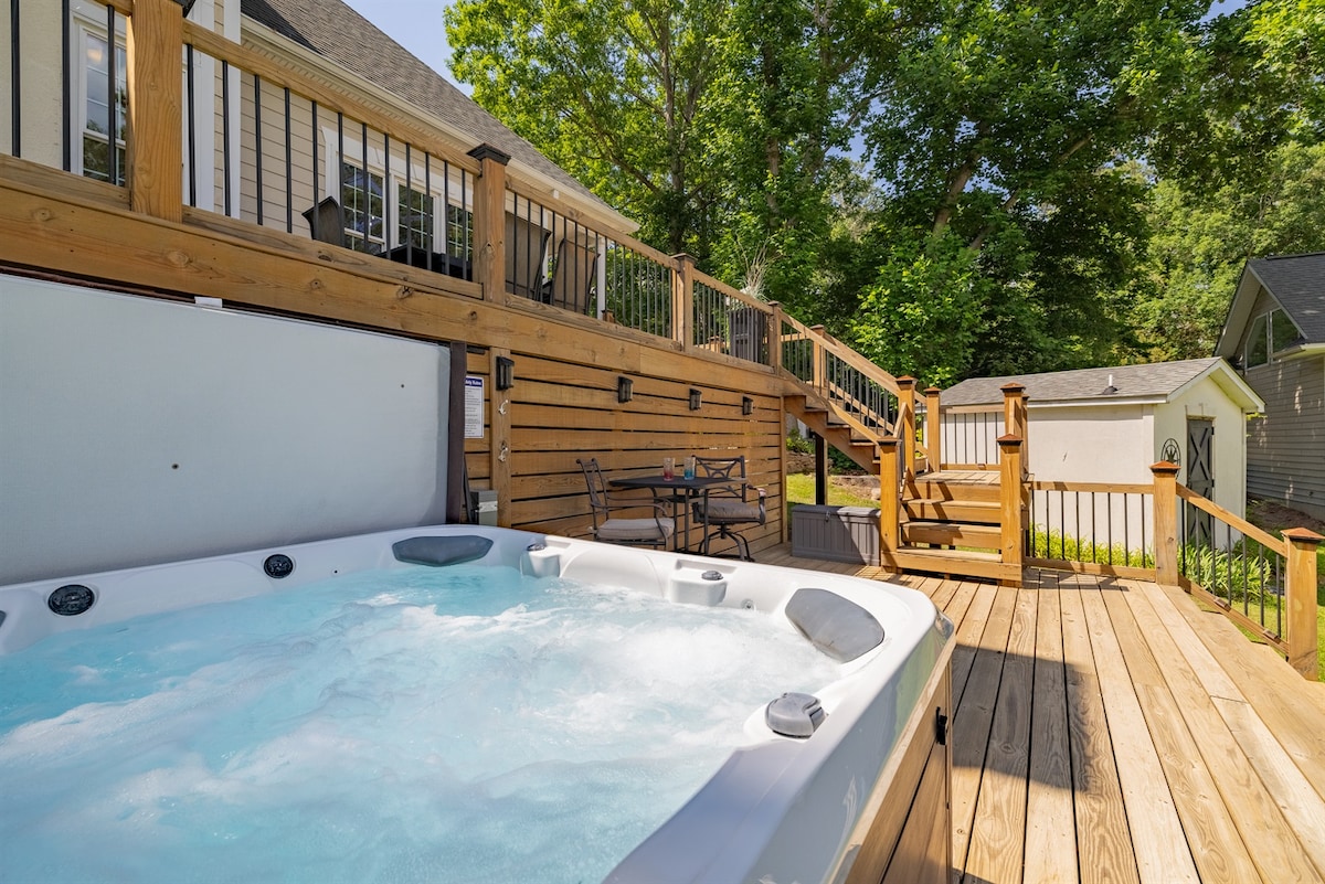 A hot tub is situated on a wooden two-tier deck, surrounded by wooden railings and green trees. A table with seating is also visible nearby, along with steps leading to the upper level of the deck. Natural light fills the space, enhancing its inviting feel.