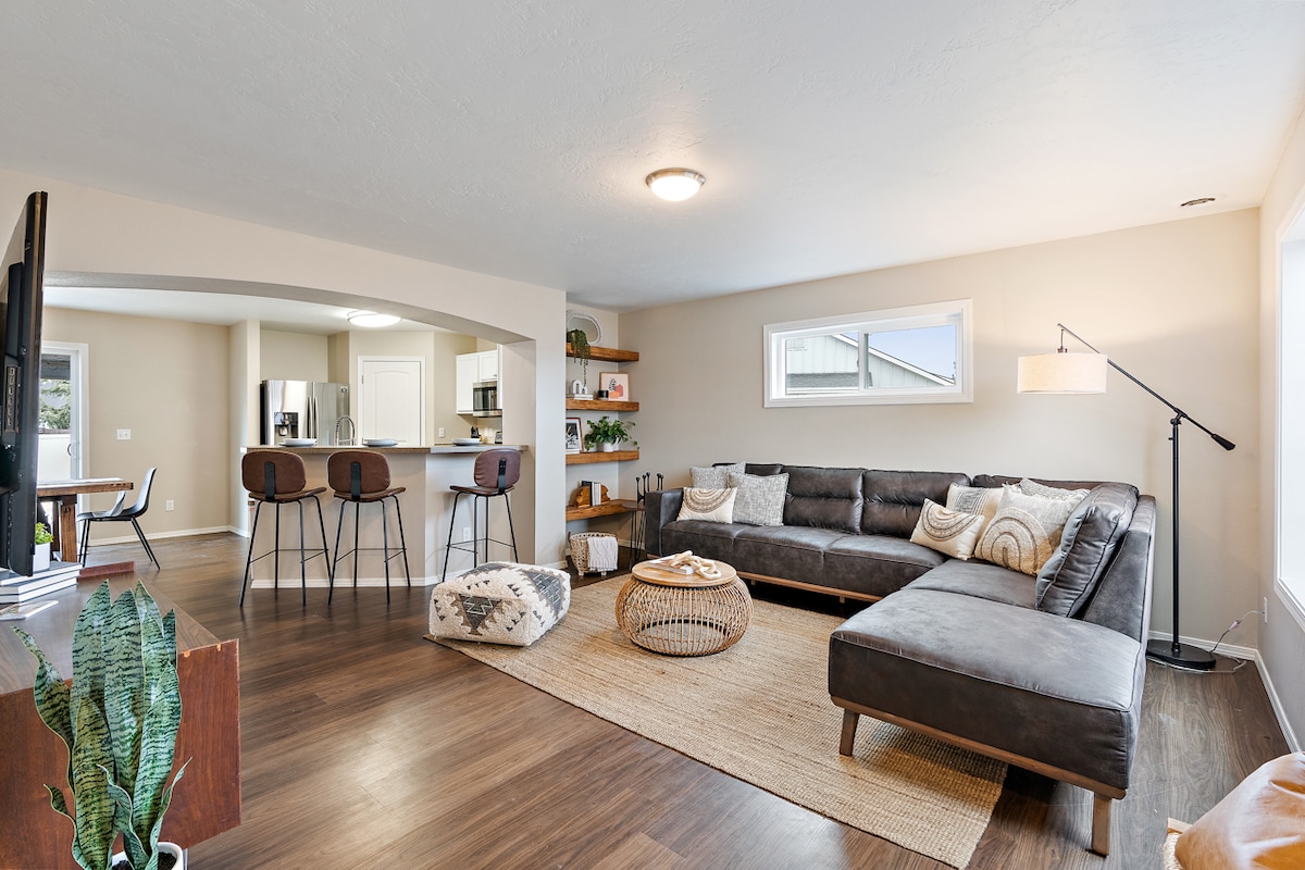 A spacious living area features a comfortable sectional sofa arranged around a woven area rug. Natural light enters through large windows, illuminating minimalist decor and modern shelving. A dining table with chairs is visible in the background, along with stylish bar stools at a kitchen counter.