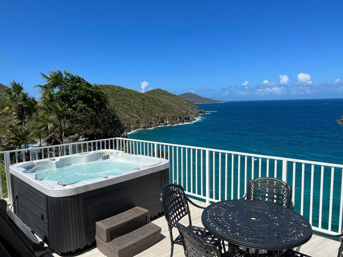 A hot tub is positioned on a deck overlooking a stunning ocean view. Black outdoor furniture is visible nearby, creating a space for relaxation. Lush greenery and gentle hills frame the scene, with clear blue skies and turquoise waters enhancing the tranquil atmosphere.