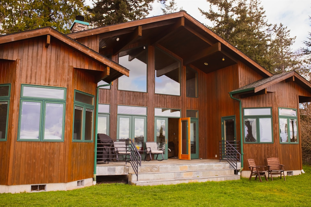 The exterior of the house showcases a mix of natural wood and large windows, allowing for ample natural light. The entrance features a bright orange door, complemented by a spacious outdoor area with chairs and a well-maintained lawn. Surrounding trees provide a serene backdrop.