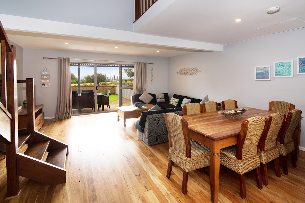 The living area features a warm wood floor and comfortable seating arrangement, with a large dining table and woven chairs. Natural light filters in through glass sliding doors, offering a view of the outdoor space. Neutral tones are complemented by coastal-themed artwork on the walls.