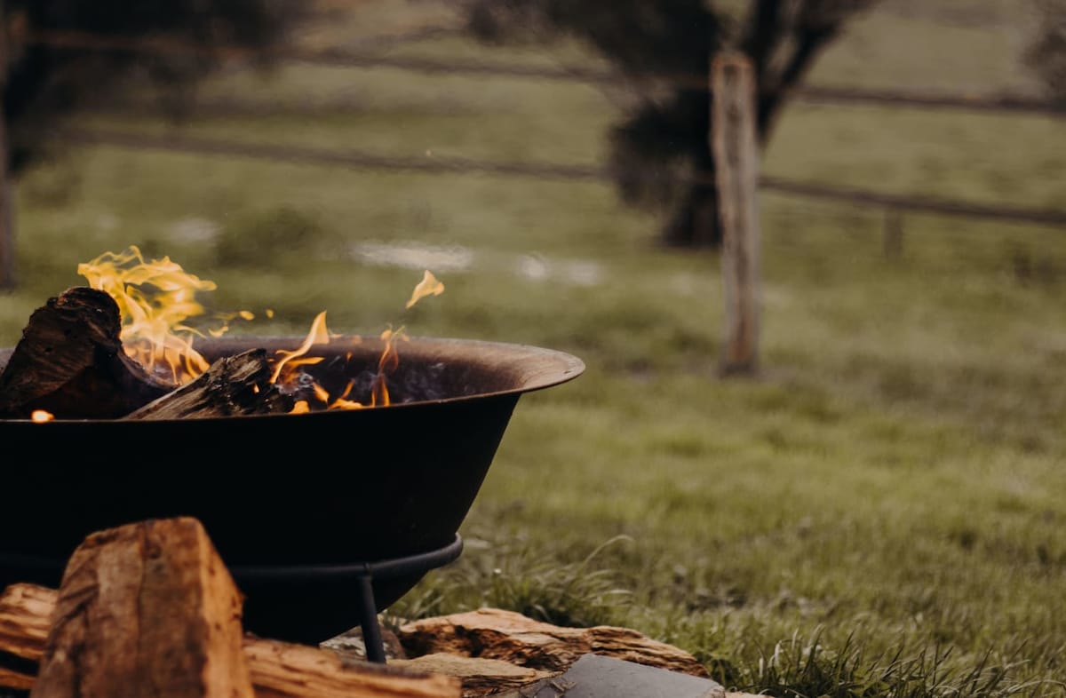 A firepit is seen glowing with flames, surrounded by logs. The grassy landscape in the background is framed by a wooden fence, creating a serene outdoor setting.