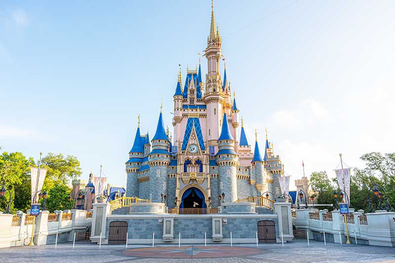 A grand castle structure showcases magnificent spires and turrets accented with blue roofs and gold details. Surrounding gardens add lush greenery, while pathways and decorative banners enhance the welcoming ambiance leading to the castle's main entrance.