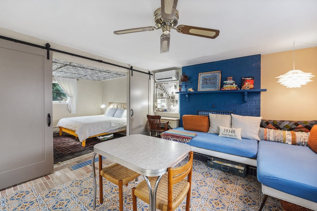 An open living area features a small dining table surrounded by chairs, adjacent to a comfortable corner sofa. Sliding barn doors lead to a bedroom with a spacious bed. A ceiling fan and unique wall art contribute to the inviting ambiance.