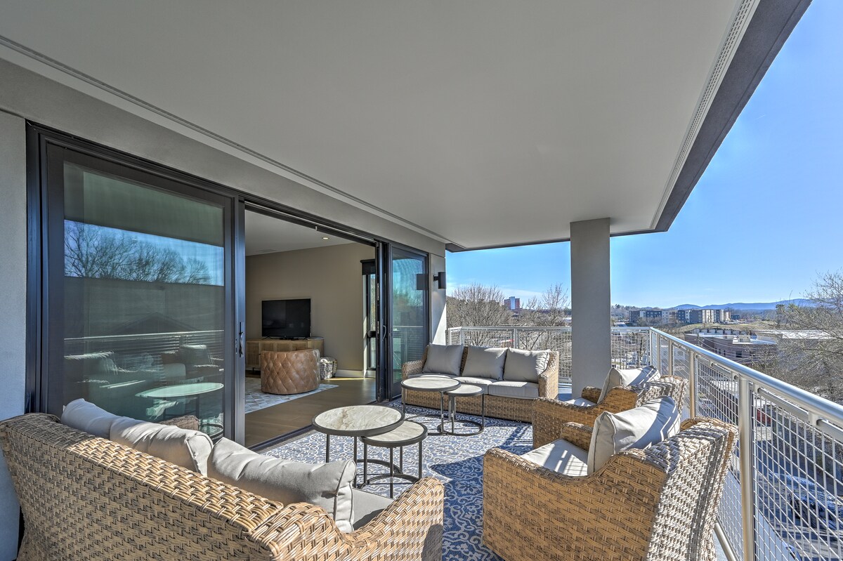 A spacious balcony features comfortable seating with woven furniture arranged around a circular table. Floor-to-ceiling glass doors provide access to the indoor living area, allowing natural light to illuminate the space. The surrounding views include distant mountains and the skyline of downtown Asheville.