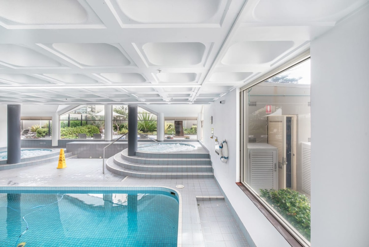 The indoor heated pool area features a tiled pool with a gentle slope leading to a shallow section. Surrounding greenery is visible through large windows, providing a serene view. Noticeable are the maintenance signs and clean, well-maintained surfaces throughout the space.