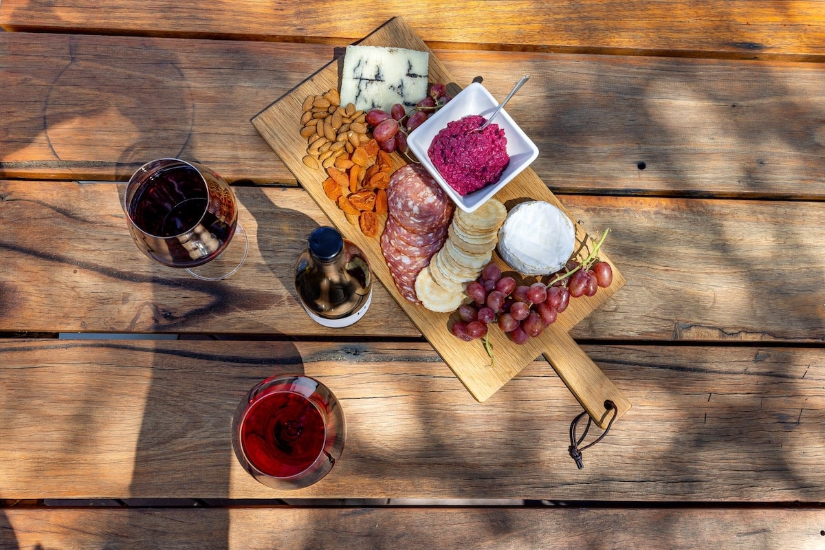 A wooden serving board displays an assortment of cheese, cured meats, and fruits, including grapes and dried apricots. A small bowl holds a vibrant spread, and two glasses of wine sit alongside. The setting suggests a relaxed outdoor dining experience.