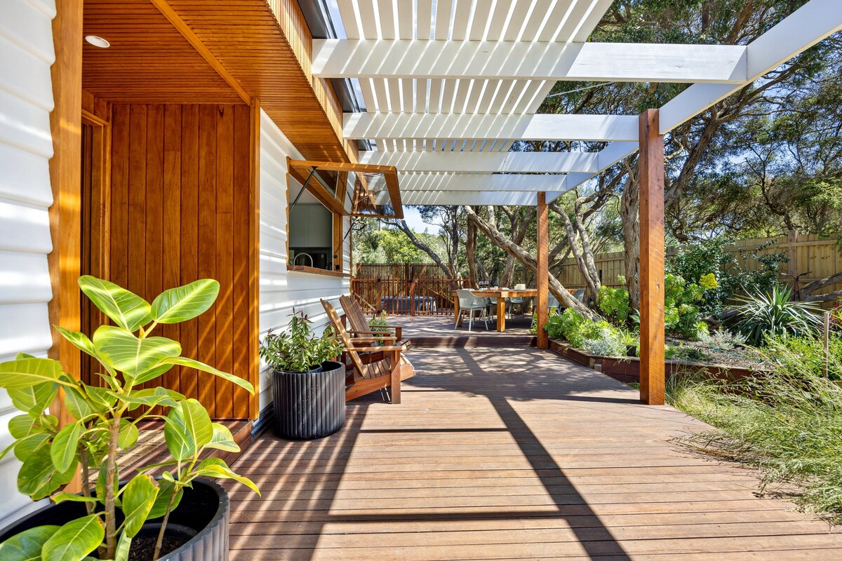 A spacious wooden deck features a mix of lounge chairs and outdoor dining options under a covered area. Lush greenery is visible nearby, with planters adding texture to the outdoor setting. Sunlight filters through the slatted roof, creating a dynamic play of light and shadow.