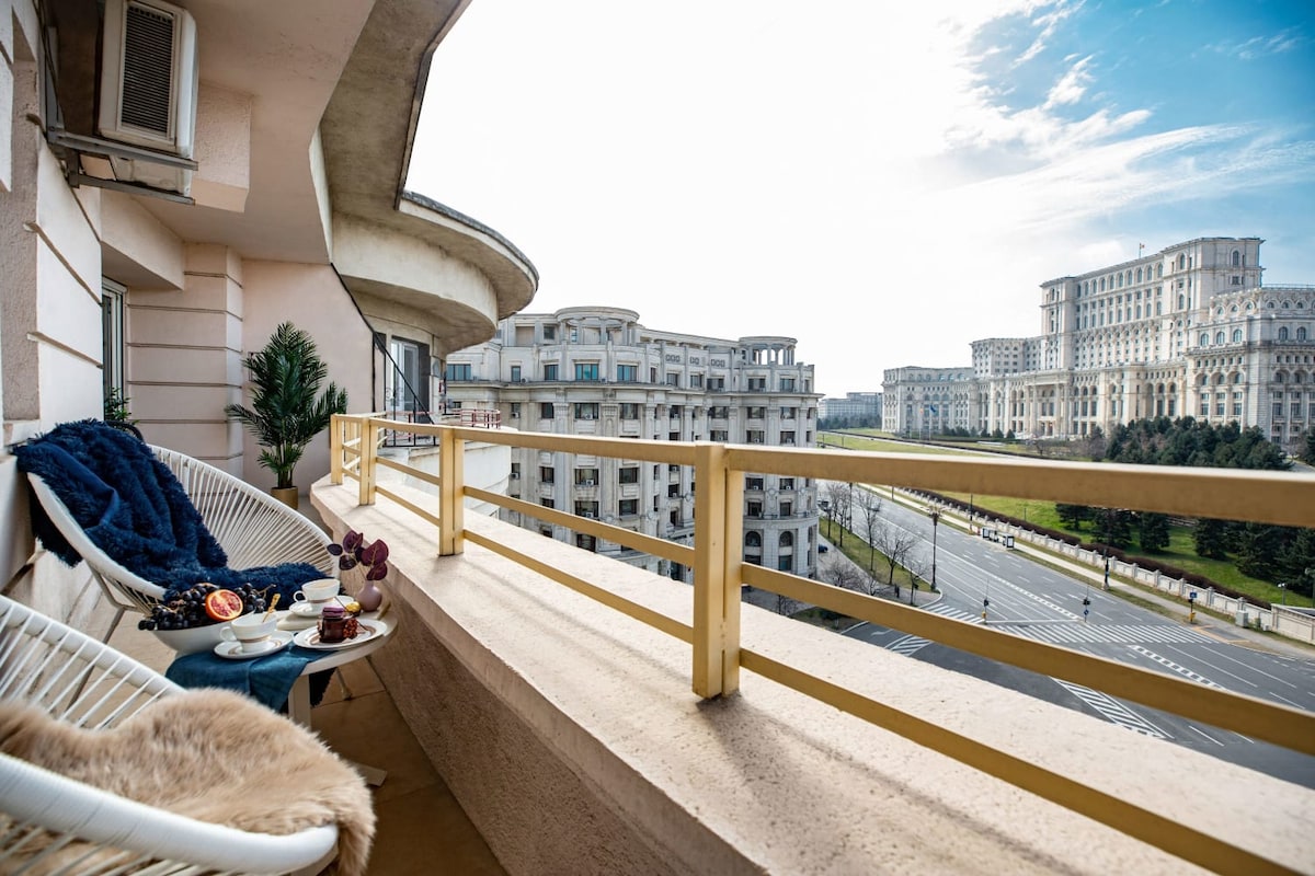 An outdoor terrace features comfortable seating alongside a small table set with coffee and snacks. The view includes the House of Parliament in the background, framed by a clear blue sky. Neighboring buildings and a tree line are visible across the street.