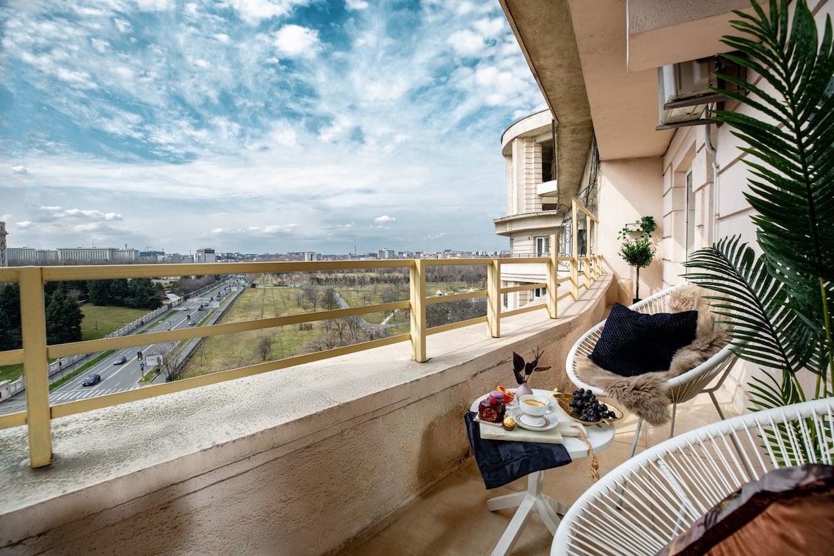 An outdoor terrace provides a serene seating area, adorned with comfortable chairs and a small table set for coffee. A panoramic view of the city skyline and greenery captivates attention under a partly cloudy sky, enhancing the relaxed atmosphere.