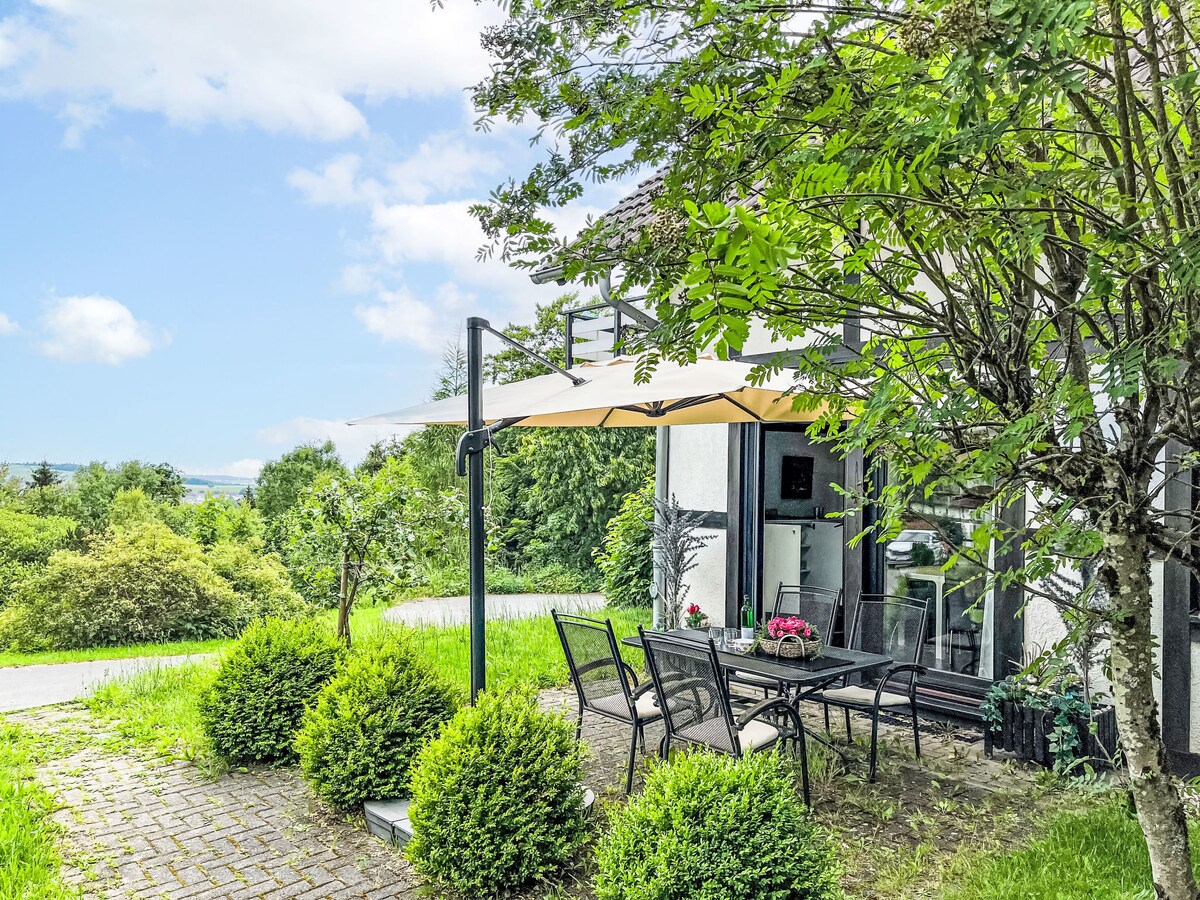 A terrace area is presented with a table and chairs under a sunshade, surrounded by lush greenery. Potted flowers are placed on the table, with a view of the tranquil landscape in the background. The ground is paved with stone tiles, enhancing the outdoor relaxation space.