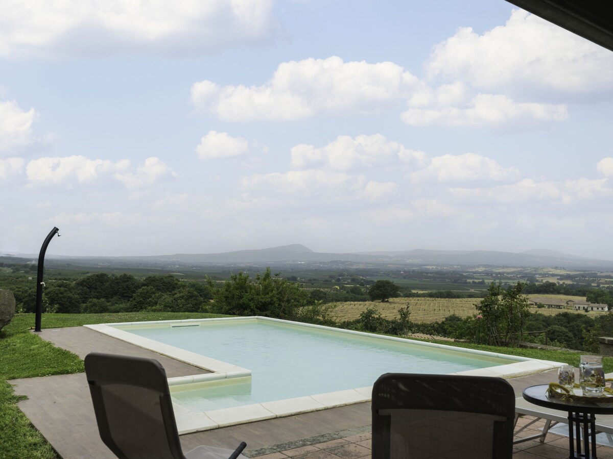 An inviting swimming pool is framed by a wooden deck, set against a vast landscape of rolling hills and distant mountains. Comfortable seating with two chairs and a table is positioned nearby, providing a perfect view of the serene surroundings under a partly cloudy sky.