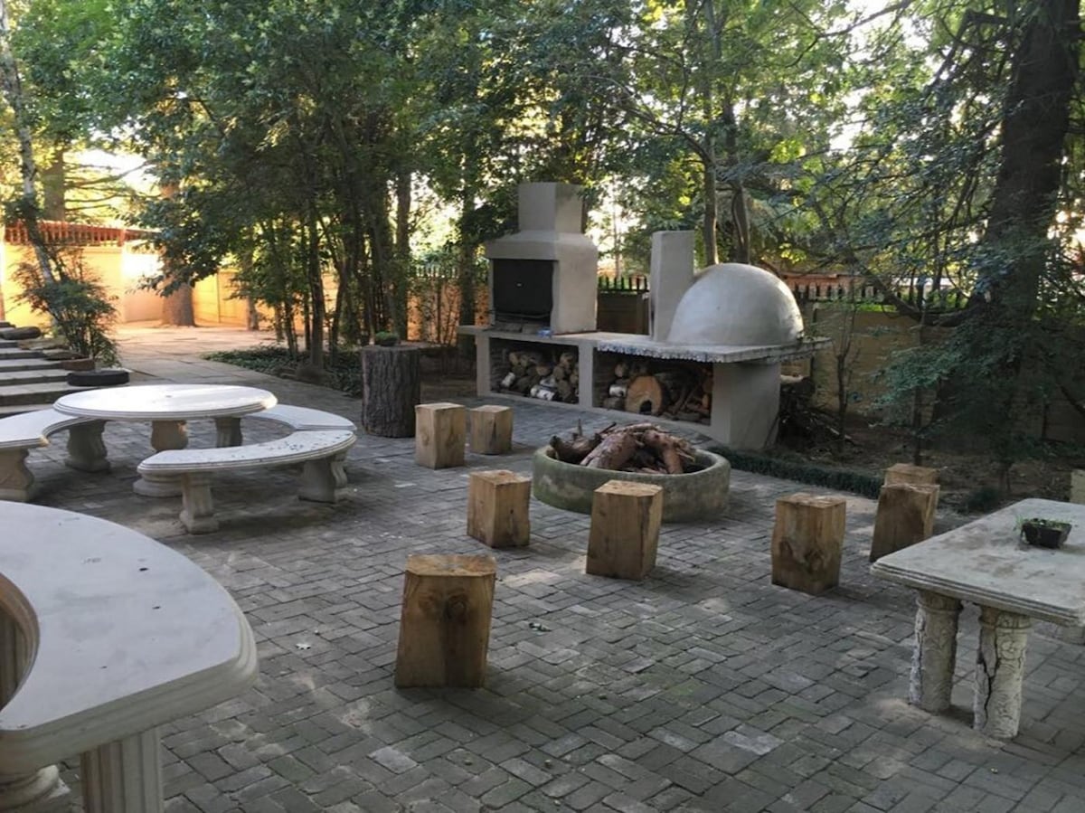An outdoor seating area is surrounded by lush trees, featuring stone benches and rustic wooden stools. A stone fire pit is situated at the center, with a barbecue grill and oven nearby. The paved surface creates a welcoming space for gatherings.