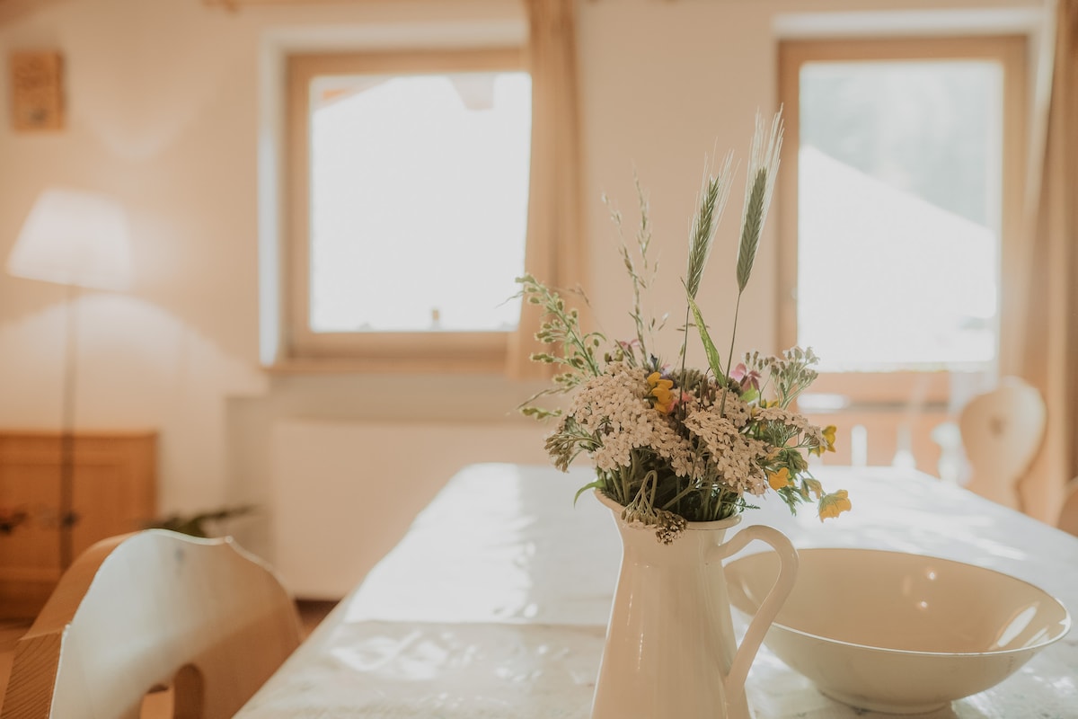 A dining area is visible, featuring a light wooden table set with a simple white bowl and a vase of fresh wildflowers. Two windows allow natural light to illuminate the space, while a soft glow emanates from a nearby floor lamp.