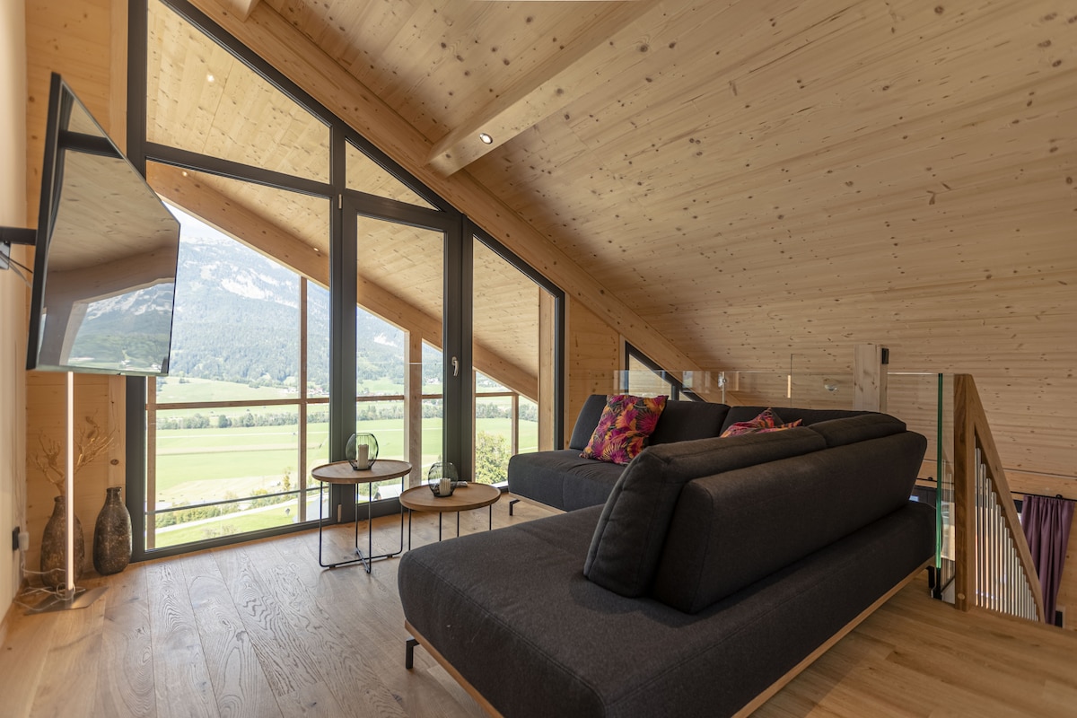 A modern loft space features large windows that provide expansive views of the surrounding landscape. Comfortable seating is arranged in a cozy configuration. Light wood paneling complements the overall design, and a flat-screen television is mounted on the wall.