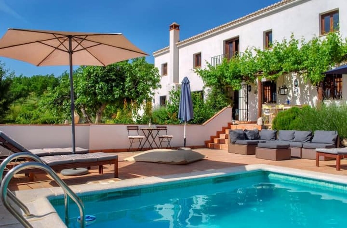 A serene outdoor area surrounds the inviting blue pool, with a sun lounger and an umbrella providing shade. Soft seating arranged around a low table offers relaxation space. A staircase leads to the entrance of the two-story house, bordered by lush greenery.