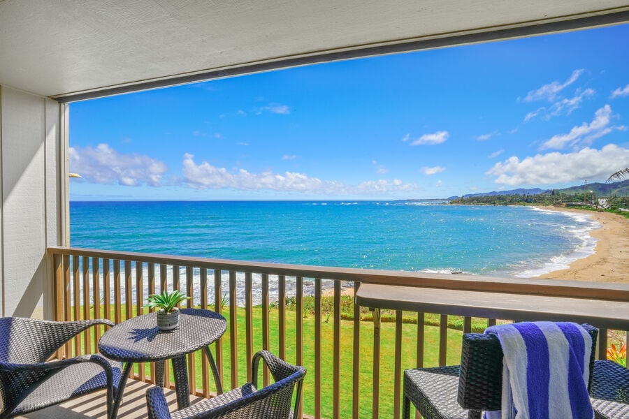 A spacious private lanai overlooks the expansive ocean, with gently crashing waves and a sandy beach visible in the distance. Two chairs and a small table are arranged on the balcony, offering a serene outdoor space to enjoy the coastal views and gentle sea breeze.