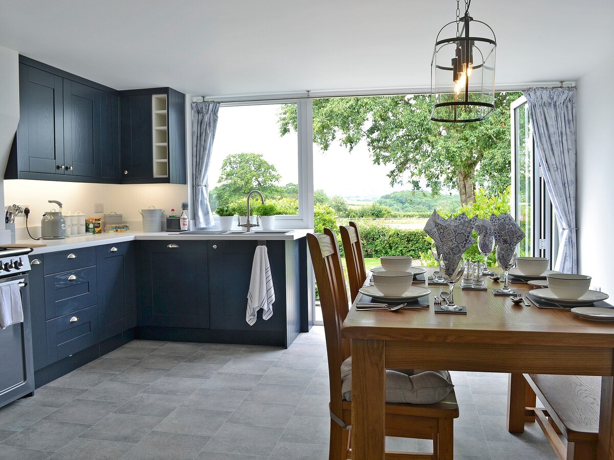 The kitchen features modern cabinetry and a spacious dining area. A large window offers scenic views of the surrounding greenery, while patio doors provide access to the outdoor space. The dining table is set with tableware, complemented by a light fixture above.