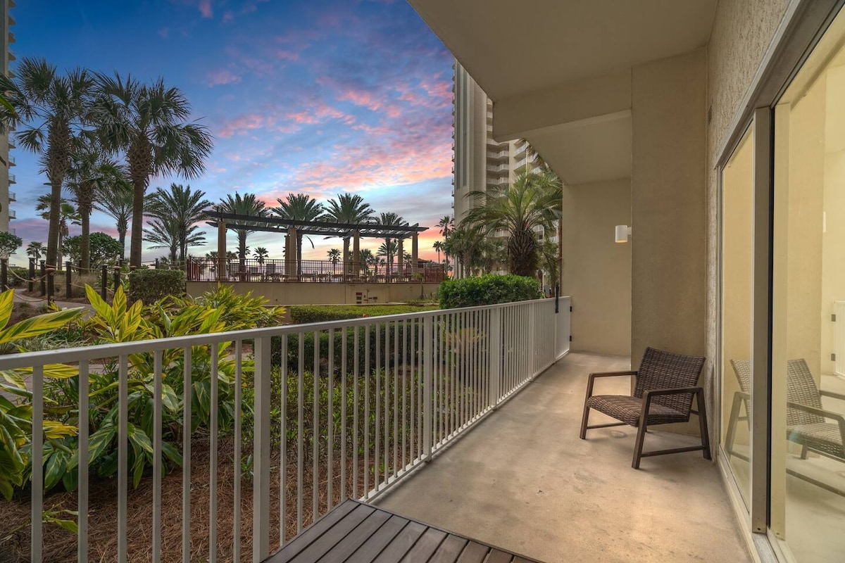 A private balcony with a comfortable chair overlooks a lush courtyard featuring palm trees and tropical plants. The horizon showcases a colorful sunset sky, enhancing the tranquil setting and providing direct access to the pool deck below.