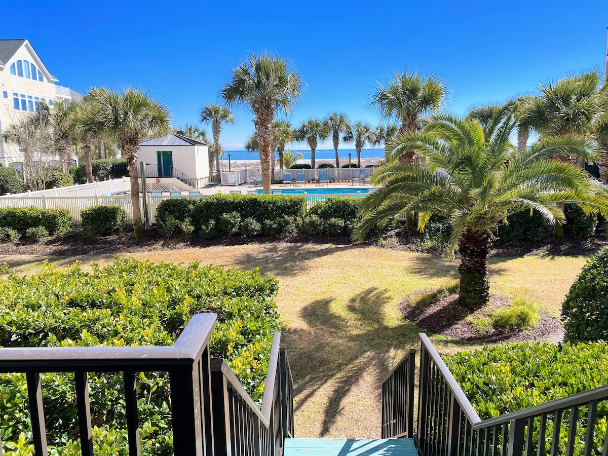 A view from a set of stairs leads onto a landscaped area featuring palm trees and manicured greenery. A pool is visible in the distance, accompanied by beach access framed by sunny blue skies.
