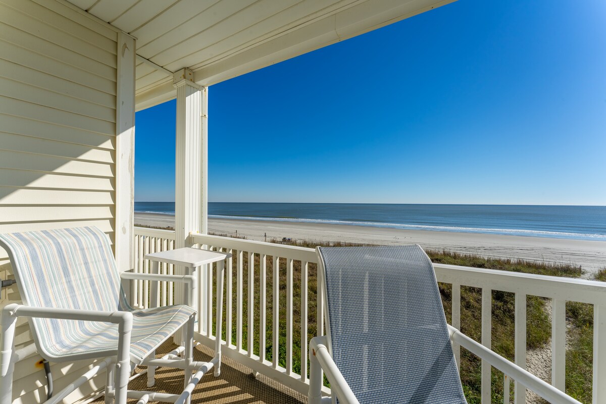 A private balcony features two lounge chairs facing the ocean, with a small side table positioned between them. The expansive view includes a sandy beach and calm waters under a clear blue sky.