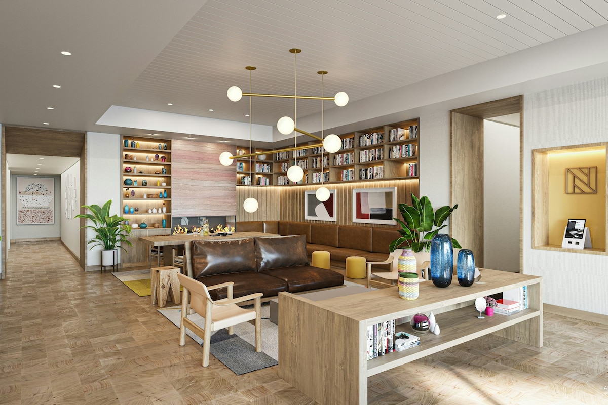 A modern lobby area features natural wood finishes and a unique light fixture with round bulbs. Comfortable seating includes a brown leather sofa and two light-colored chairs. A bookshelf displays various items, while decorative plants and colorful vases add warmth to the inviting space.