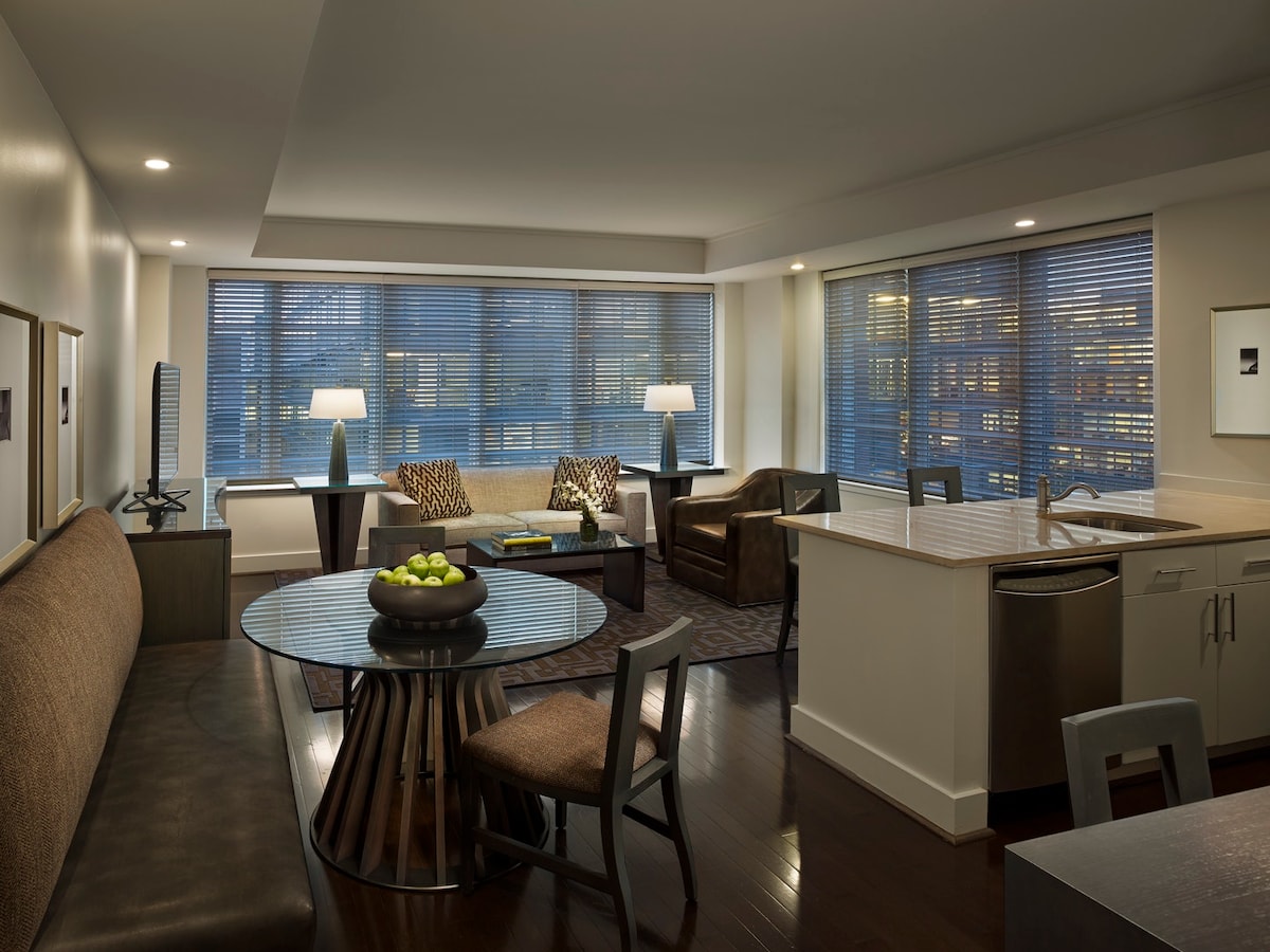 A spacious living area features a round dining table at its center, surrounded by chairs. A soft sectional couch and two armchairs provide seating, with a television placed on an adjoining wall. Natural light enters the room through large windows dressed with blinds.