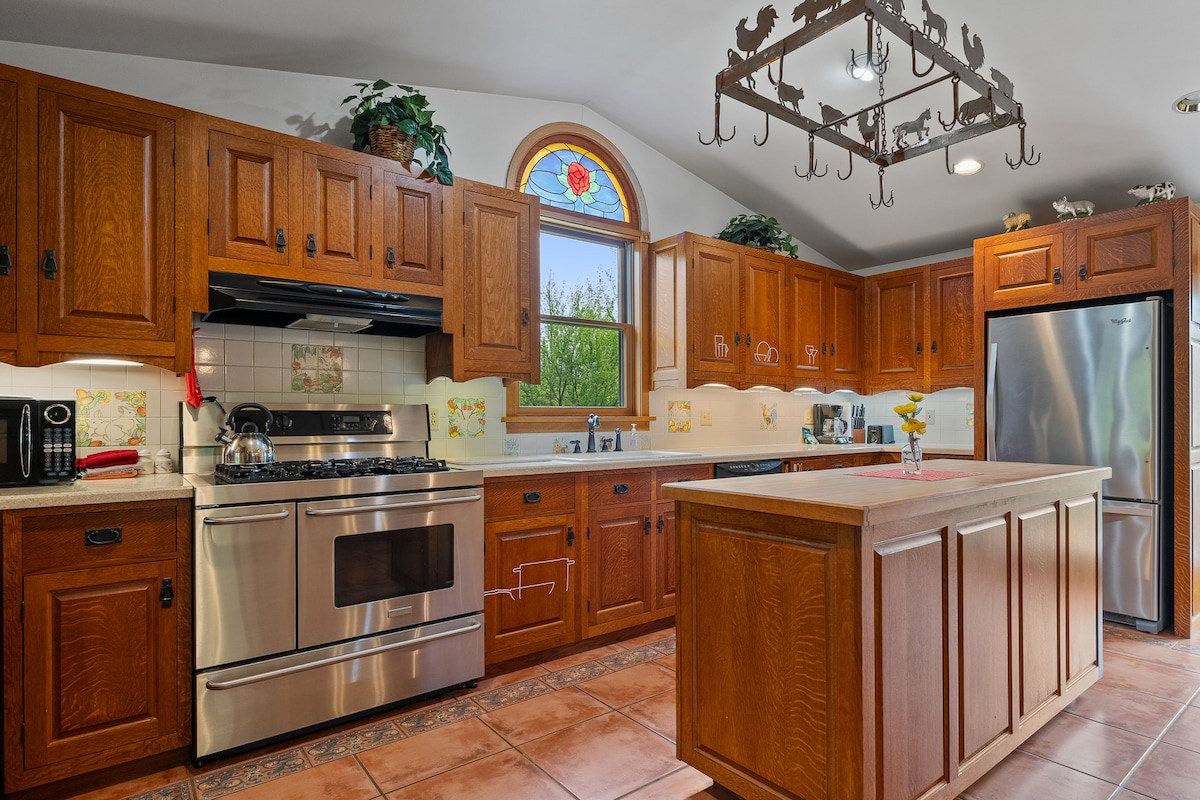 The kitchen showcases handcrafted cherrywood cabinets and a large central island with a countertop. Stainless steel appliances are present, including a gas stove and refrigerator. Natural light floods the space through a stained glass window, highlighting the warm, inviting atmosphere of the room.
