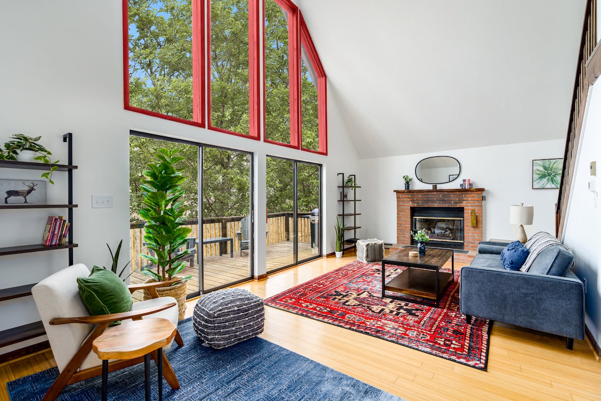 A spacious living room features large east-facing windows that allow ample natural light. A patterned area rug adds color to the wooden floor. Seating includes a cozy blue sofa, a wooden chair, and a pouf. A decorative fireplace complements the modern decor.