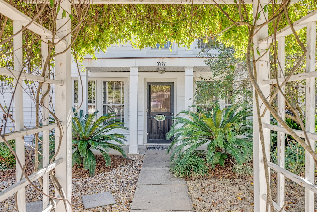 A welcoming entrance is framed by a white wooden trellis, adorned with lush green vines. Flanking the walkway, vibrant palm plants are positioned alongside the steps leading to the front door, which is centered within the classic white facade.