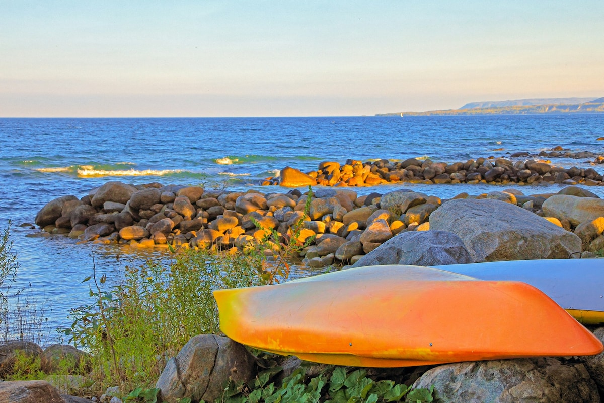 A tranquil shoreline scene features colorful kayaks resting on a rocky beach, overlooking calm blue waters. Sunlight casts a warm glow on the smooth stones, and gentle waves can be seen in the distance. The shoreline is lined with scattered greenery, enhancing the natural setting.