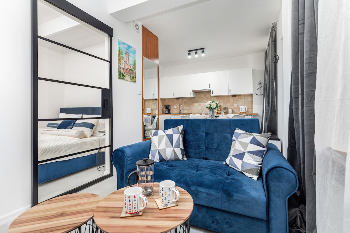 A cozy living space features a blue sofa adorned with decorative pillows, accompanied by two wooden coffee tables. A kitchenette with modern appliances is visible in the background, separated by a glass partition. Light colors dominate the decor, contributing to a bright atmosphere.