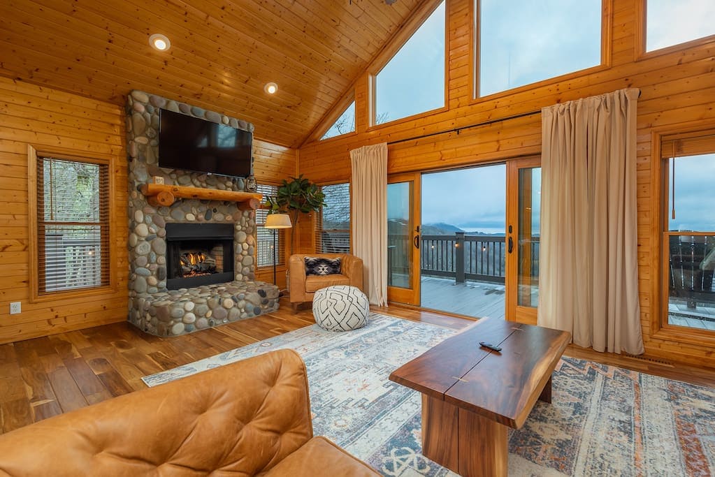 A spacious living area features a stone fireplace flanked by tall windows, offering expansive mountain views. Wooden walls and ceiling create a warm ambiance. A comfortable seating arrangement includes a tan sofa and a textured pouf, complemented by a rustic wooden coffee table and area rug.