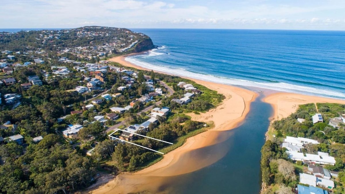 An aerial view showcases the coastline of Copacabana, with the ocean waves gently breaking on the sandy beach. Lush greenery lines the shoreline, while residential homes are visible along the riverbank, leading to the lagoon. The distant hills add depth to the scenic landscape.