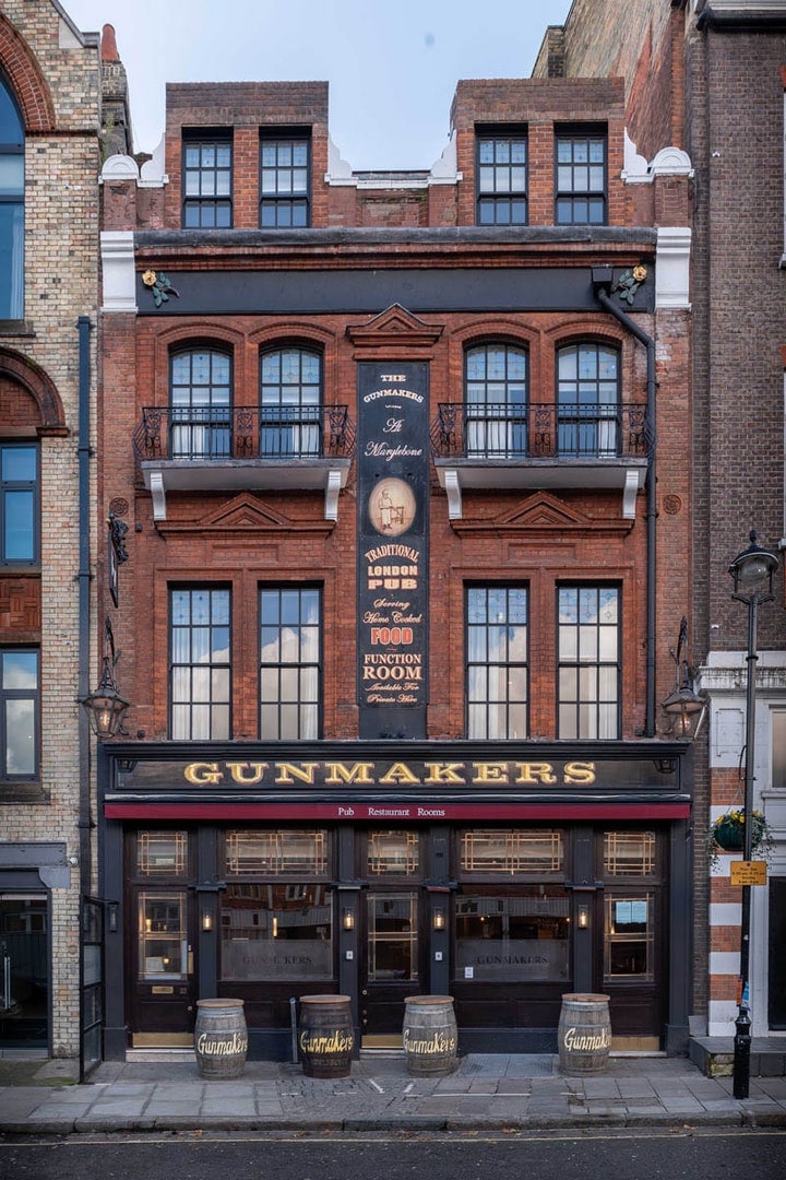 The exterior of a historic building is displayed, featuring a two-story facade with large windows and a prominent sign that reads 'GUNMAKERS.' Decorative brickwork enhances the structure, while the ground floor features wooden accents and barrels, creating a classic urban charm.