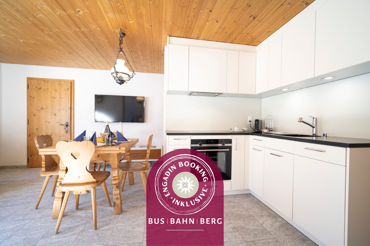 The kitchen area features modern cabinetry in neutral tones, complemented by a wooden ceiling. A dining table with wooden chairs is visible, alongside a flat-screen television. Appliances include an oven and a stovetop, providing a fully equipped cooking space.