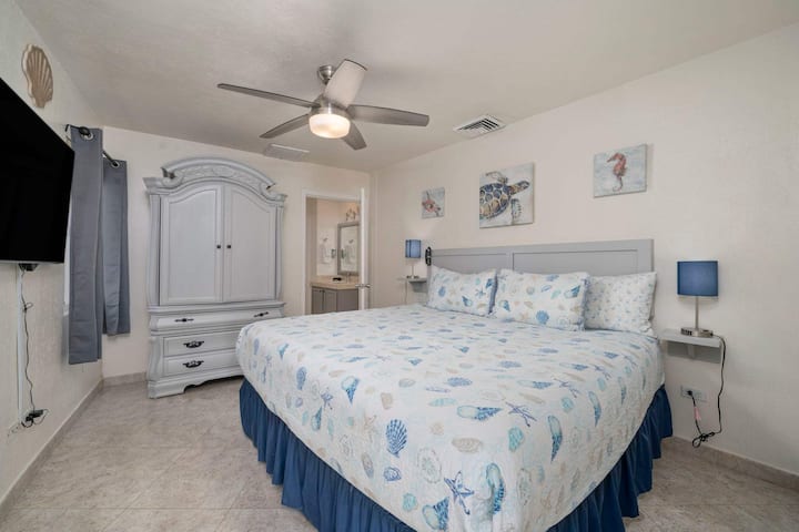 Amplio dormitorio tamaño king con TV grande, ventilador de techo y elegantes muebles costeros para una estadía tranquila.