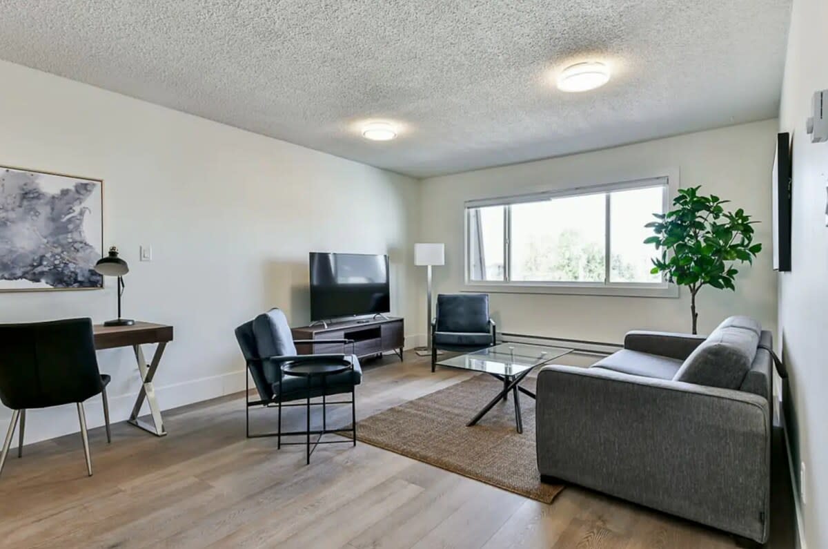 An open plan living area is presented, featuring a comfortable sofa and two chairs arranged around a glass coffee table. A flat-screen TV is positioned on a low cabinet, and a desk with a chair is located to the left. Natural light enters through a large window.