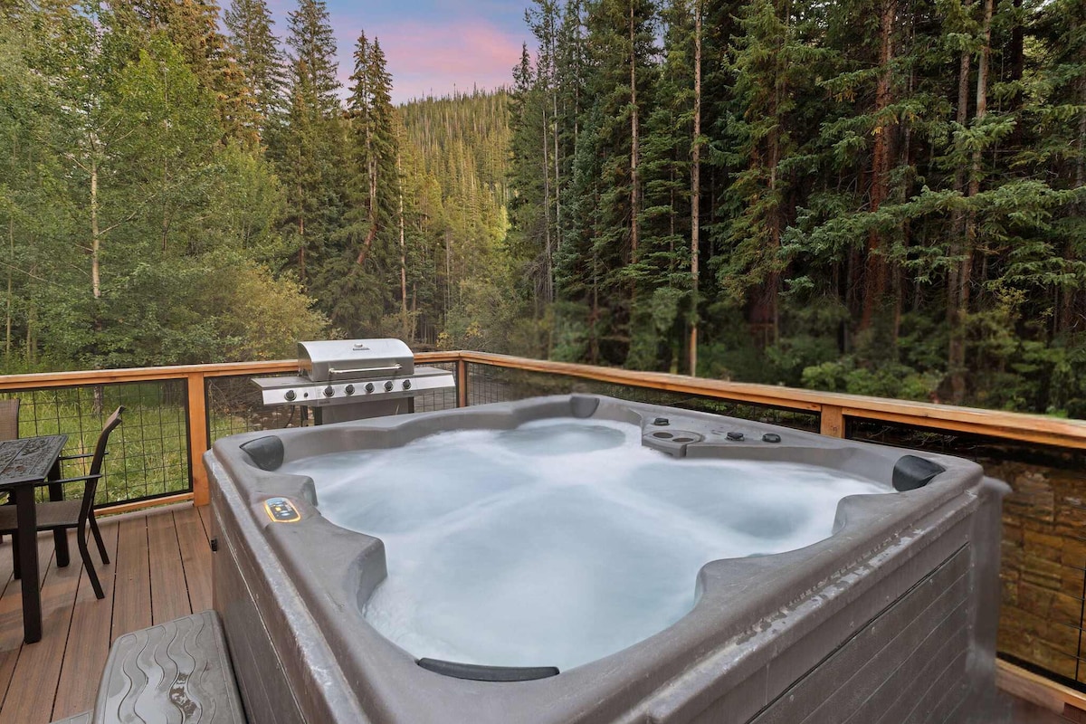 A spacious back deck is featured, showcasing a large hot tub surrounded by towering trees. A propane grill is positioned nearby, accompanied by a dining table, creating an inviting outdoor space for relaxation and mountain views.