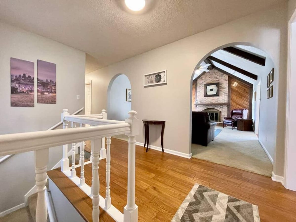 The entryway features a well-lit hallway with hardwood flooring and curved archways leading to a cozy living area. Two framed photographs hang on the wall, and a small table is positioned by the staircase. A textured area rug adds warmth to the space.