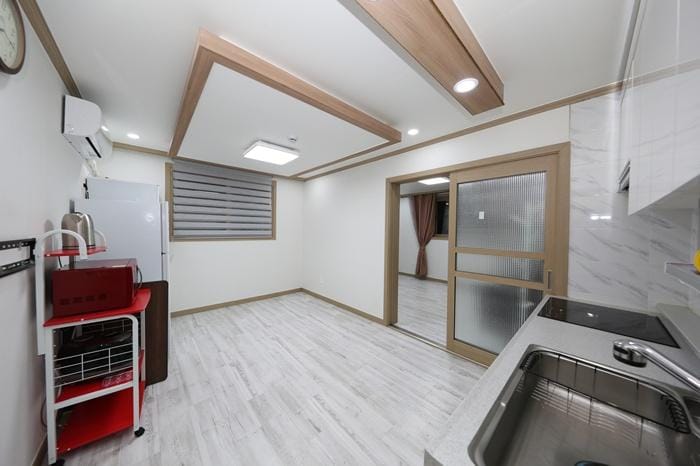 A modern kitchen area features light wood accents and a spacious layout. A sleek countertop with a sink and stove is complemented by a light-colored cabinet. A refrigerator and a cart provide additional storage. A glass door leads to another room, enhancing the openness of the space.