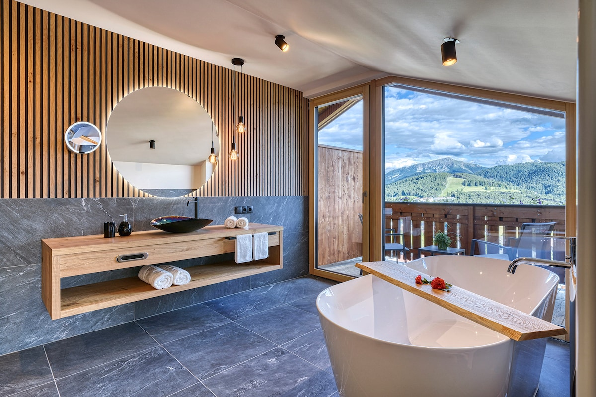 An exclusive bathroom features a free-standing bathtub and a Murano glass sink set against a backdrop of natural wood paneling. Large windows provide a view of the surrounding mountains, while a wooden shelf holds neatly arranged towels.