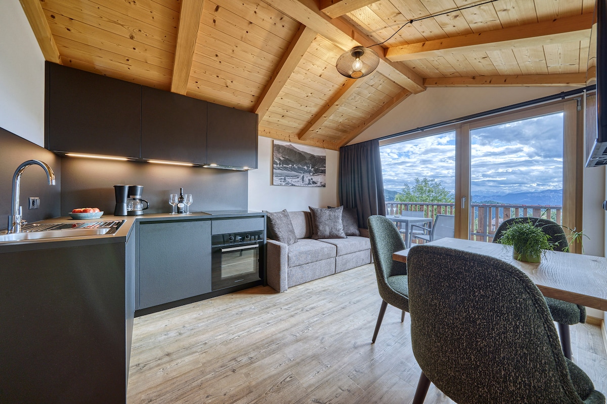 The open living area features a modern kitchen with a sleek design, a comfortable seating area with a sofa, and a dining table. Large windows frame stunning views of the mountainous landscape, enhancing the bright and airy feel of the space.