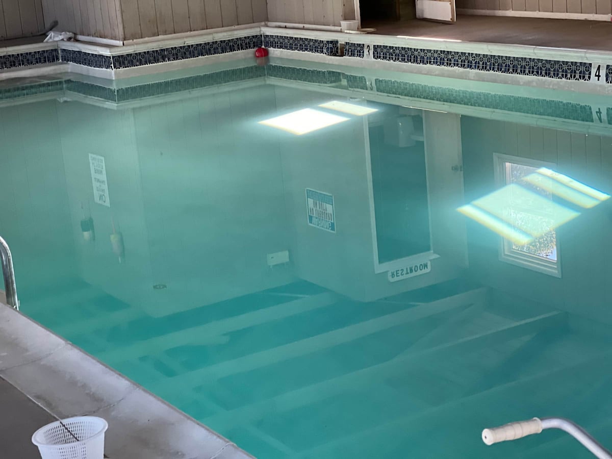 An indoor pool is featured, with clear turquoise water reflecting the overhead lights. The pool area is surrounded by a tiled border, and a nearby door offers access to additional spaces. The serene environment is designed for relaxation and enjoyment.